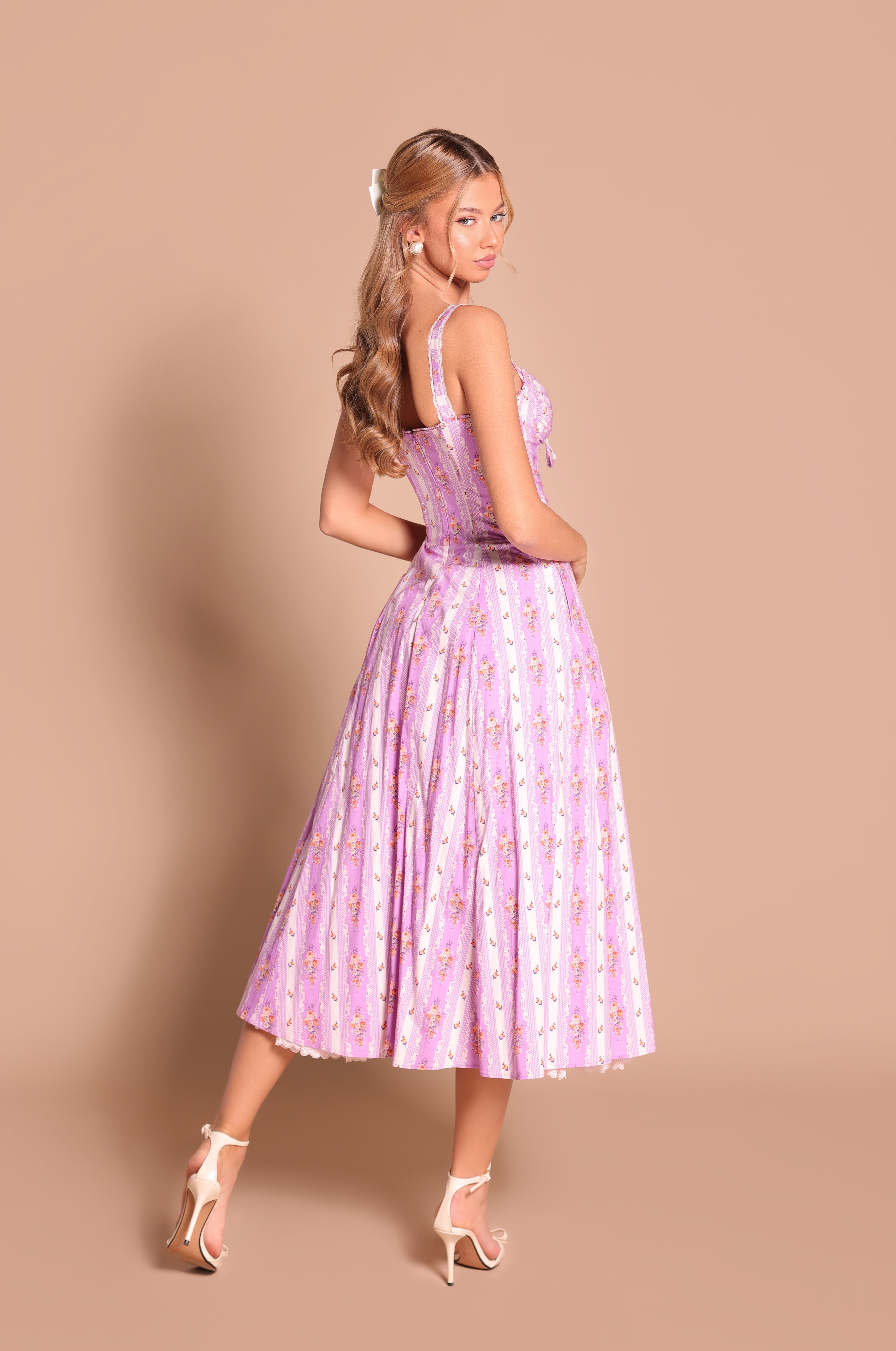 A woman in the Lilac Floral Casual Midi Dress: Celenia and white high-heeled sandals stands on a beige background, facing slightly away. Ideal for summer weddings or any sunny occasion.